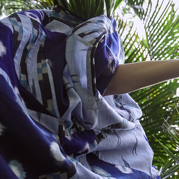 Figure in movement wearing flowing blue dress amongst green Palm leaves